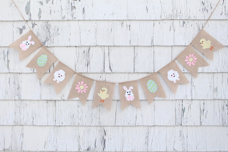 K&ouml;nnte beinhalten: Ein Osterbanner aus Jute mit einer Reihe von Flaggen, die verschiedene Ostermotive zeigen, darunter ein H&auml;schen, ein K&uuml;ken, ein Ei, eine Blume und ein Lamm. Das Banner h&auml;ngt vor einem wei&szlig;en Holz Hintergrund.