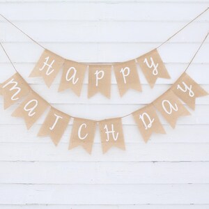 May include: Two burlap banners with white lettering spelling "Happy Match Day" are suspended against a white wooden background. The banners are strung on twine, creating a festive and celebratory atmosphere.