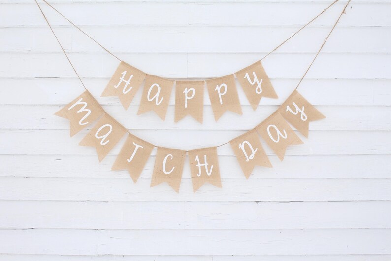 May include: Two burlap banners with white lettering spelling "Happy Match Day" are suspended against a white wooden background. The banners are strung on twine, creating a festive and celebratory atmosphere.