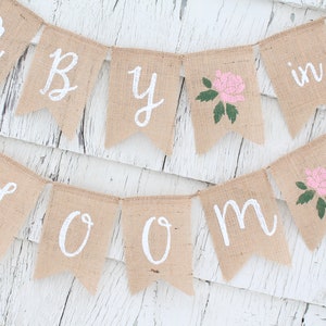 May include: A burlap banner with the words "Baby in Bloom" in white paint. The banner has pink roses with green leaves painted on it.