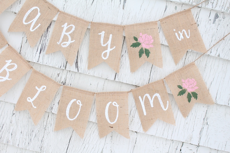 May include: A burlap banner with the words "Baby in Bloom" in white paint. The banner has pink roses with green leaves painted on it.