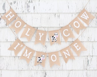 May include: A burlap banner with the text "Holy Cow I'm One" in white letters. The banner has two black and white cow illustrations.
