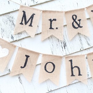 K&ouml;nnte beinhalten: Ein Banner aus Jute mit den Worten "Mr. & Mrs. John" in schwarzer Schrift. Das Banner hat ein wei&szlig;es Herz auf einem der Fahnen.