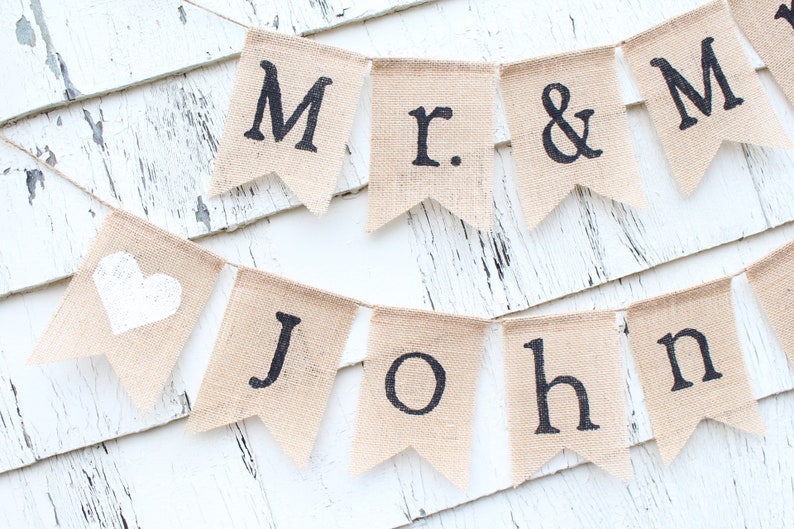 K&ouml;nnte beinhalten: Ein Banner aus Jute mit den Worten "Mr. & Mrs. John" in schwarzer Schrift. Das Banner hat ein wei&szlig;es Herz auf einem der Fahnen.