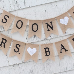 May include: A burlap banner with the words "MRS. SOON TO BE BARR" in black lettering. The banner has two white hearts and is strung on a white wall.