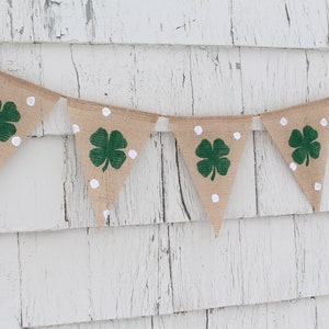 May include: A burlap banner with triangular flags, each adorned with a green shamrock and white polka dots. The banner is strung against a weathered, white-painted wooden background, perfect for St. Patrick's Day decorations.