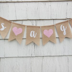 May include: It's a girl burlap banner with pink hearts and white lettering.