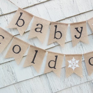 May include: A burlap banner with the words "baby cold out" in black lettering. The banner is decorated with white snowflakes.