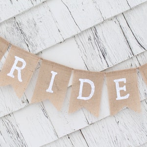 May include: A burlap banner with the word "BRIDE" in white paint and a blue starfish on the last flag. The banner is hanging on a white wooden wall.