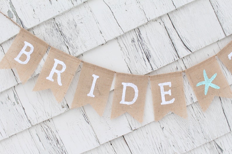 May include: A burlap banner with the word "BRIDE" in white paint and a blue starfish on the last flag. The banner is hanging on a white wooden wall.