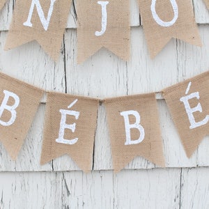 May include: A burlap banner with the words "B&eacute;b&eacute;" in white paint. The banner is strung up on a white wooden wall. The banner is decorated with pink Eiffel Towers.