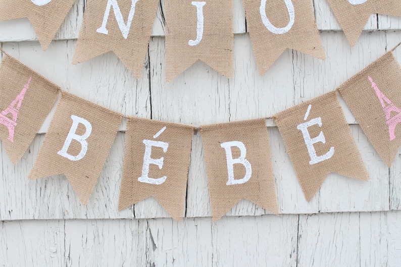 May include: A burlap banner with the words "B&eacute;b&eacute;" in white paint. The banner is strung up on a white wooden wall. The banner is decorated with pink Eiffel Towers.
