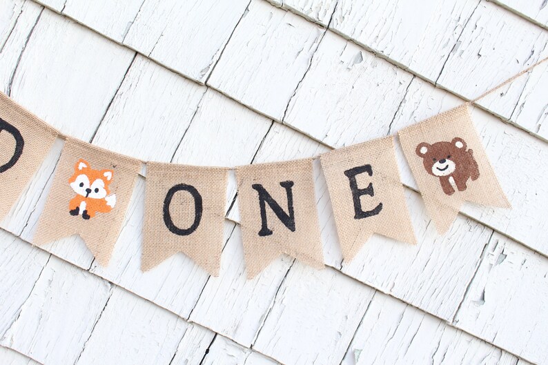 May include: A burlap banner with the word "ONE" in black letters. The banner is decorated with a fox and a bear.
