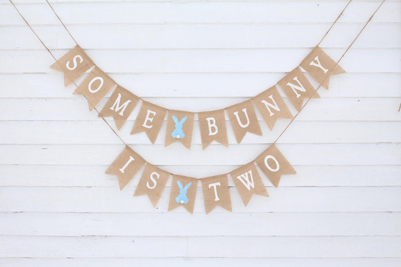 May include: A burlap banner with the text "Some Bunny Is Two" in white letters. The banner is decorated with blue bunny cutouts.