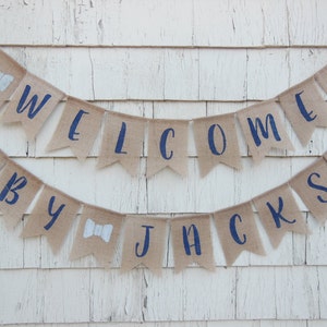 May include: A burlap banner with navy blue lettering that reads "Welcome Baby Jackson". The banner is decorated with white bowties.