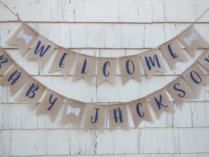 May include: A burlap banner with navy blue lettering that reads "Welcome Baby Jackson". The banner is decorated with white bowties.