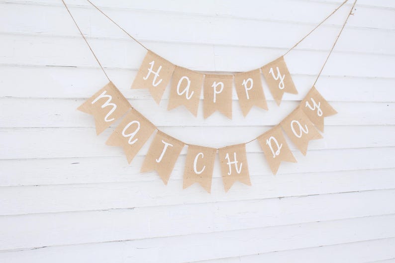 May include: A banner with the words "Happy Match Day" written in white script on individual tan pennants. The banner is strung on twine and hangs against a white wooden background.