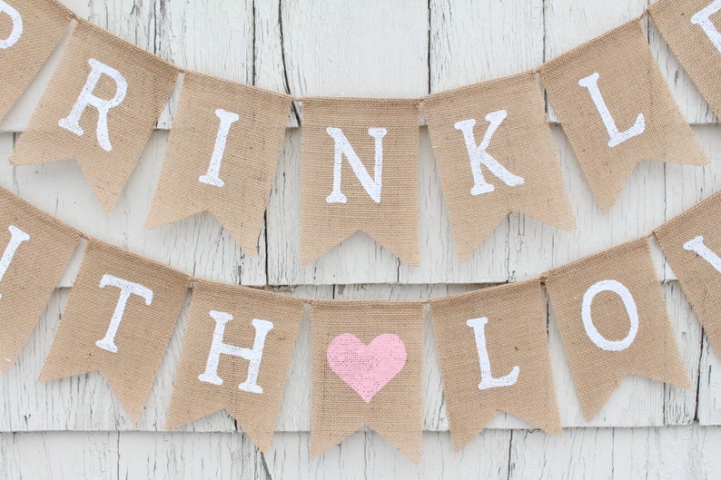 May include: A burlap banner with the words "Sprinkle with Love" in white lettering. A pink heart is in the middle of the banner.