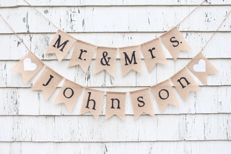 K&ouml;nnte beinhalten: Ein Banner aus Jute mit wei&szlig;en Herzen und schwarzem Text, der "Mr. & Mrs. Johnson" lautet.