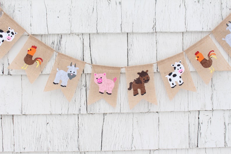 May include: A burlap banner with farm animal illustrations. The banner features a cow, rooster, goat, pig, horse, and another cow. The banner is strung on a white wooden wall.