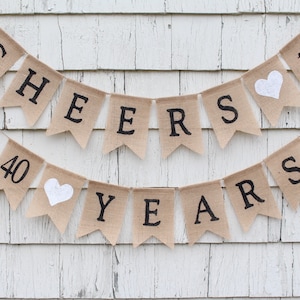 Könnte beinhalten: Ein Banner aus Jute mit dem Text "Cheers to 40 Years" in schwarzen Buchstaben. Das Banner hat weiße Herzen zwischen den Wörtern.