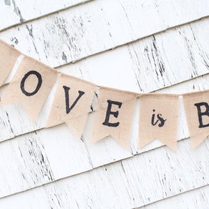 May include: A burlap banner with the text "LOVE is BR" hanging on a white wooden wall.
