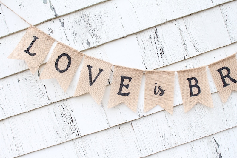 May include: A burlap banner with the text "LOVE is BR" hanging on a white wooden wall.