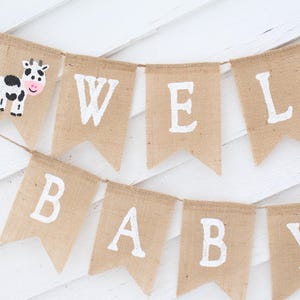 Pode incluir: Uma faixa de serapilheira com as palavras "WELCOME BABY" em letras brancas. Uma bandeira apresenta uma ilustra&ccedil;&atilde;o de vaca. A faixa &eacute; pendurada em uma corda em um fundo de madeira branca. Ideal para um ch&aacute; de beb&ecirc;.