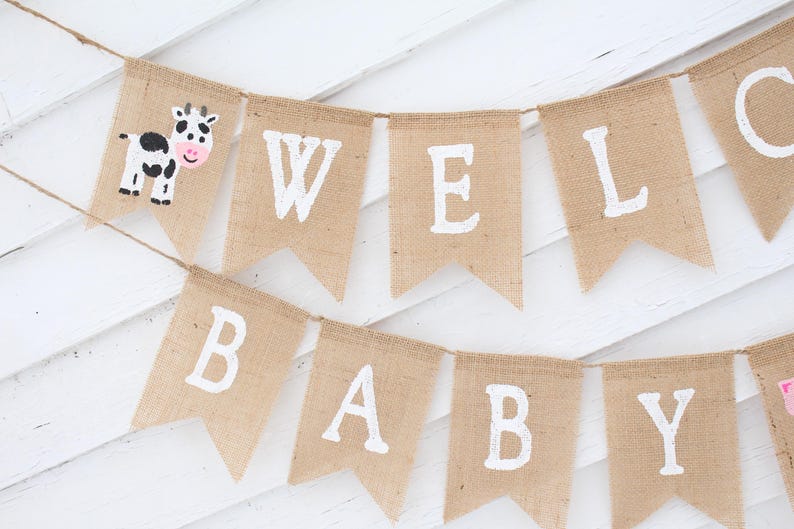 Pode incluir: Uma faixa de serapilheira com as palavras "WELCOME BABY" em letras brancas. Uma bandeira apresenta uma ilustra&ccedil;&atilde;o de vaca. A faixa &eacute; pendurada em uma corda em um fundo de madeira branca. Ideal para um ch&aacute; de beb&ecirc;.