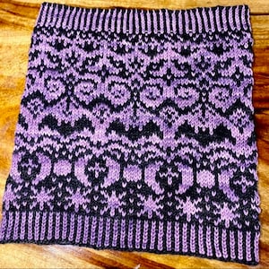 Pode incluir: Um cachecol de malha em tons de roxo e preto. O cachecol apresenta um padrão geométrico repetitivo com uma parte superior e inferior caneladas. O design inclui formas florais e em forma de coração. O cachecol é feito de um material macio e quente.