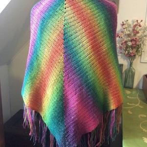 May include: A rainbow colored knitted shawl with fringe on the edges. The shawl is draped over a mannequin.
