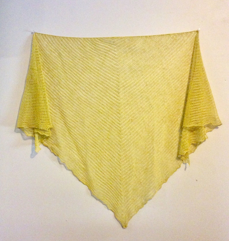May include: A yellow crocheted shawl with a chevron pattern. The shawl is hanging on a white wall.