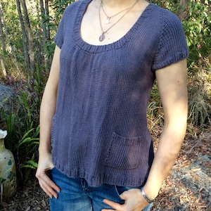 May include: A short-sleeved, indigo-coloured knitted top with a scoop neckline and a single pocket. The top is paired with blue jeans. The top has a textured appearance and a relaxed fit, suitable for casual wear.