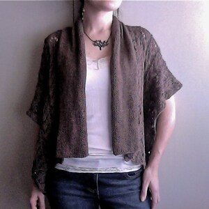 Può includere: Un cardigan marrone lavorato a uncinetto con un motivo a pizzo. Il cardigan è aperto sul davanti e ha una forma lunga e fluida. La modella indossa una canottiera bianca sotto il cardigan.