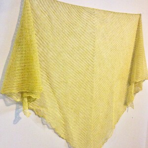 May include: A yellow knitted shawl with a delicate, open weave pattern. The shawl is draped over a white wall and has a slightly scalloped edge.