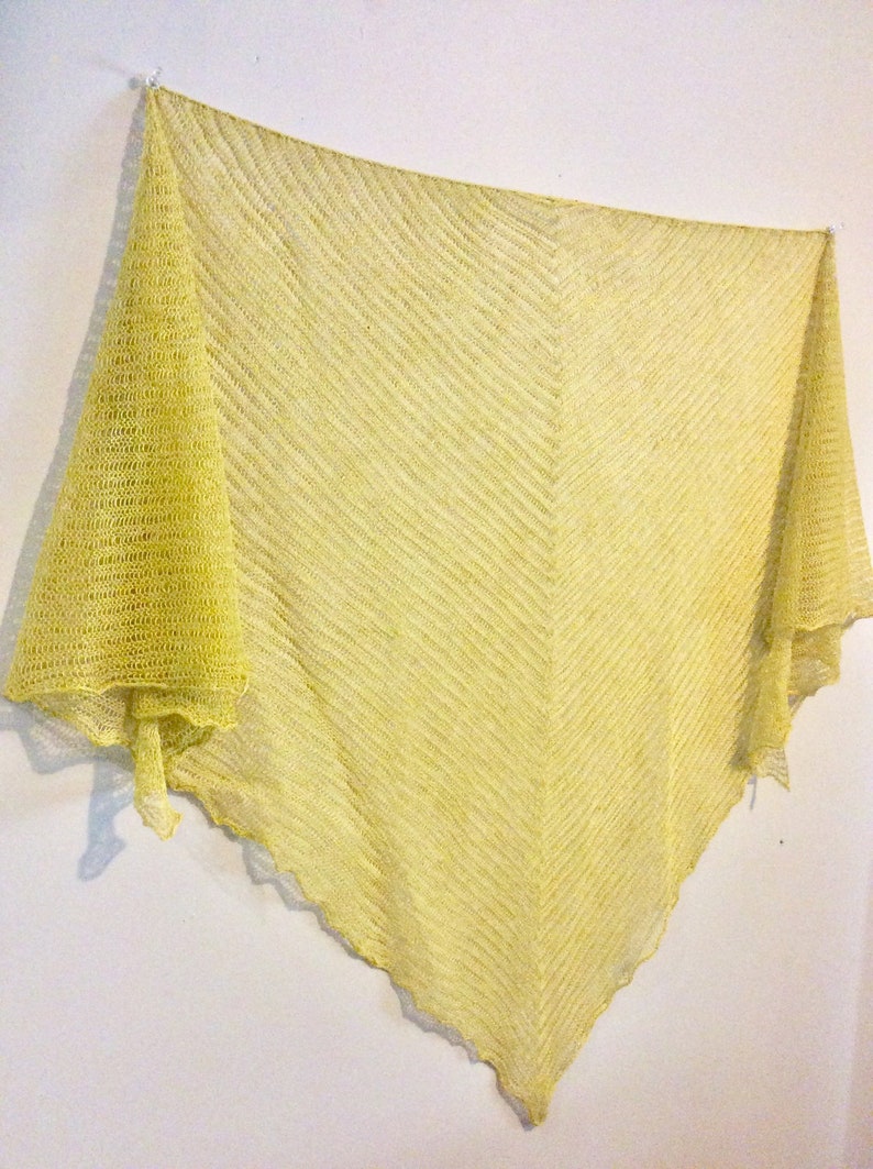 May include: A yellow knitted shawl with a delicate, open weave pattern. The shawl is draped over a white wall and has a slightly scalloped edge.