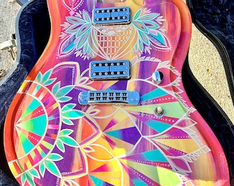 Op de afbeelding: Een op maat geschilderde elektrische gitaar met een kleurrijk mandala-ontwerp. De gitaar heeft een fel oranje, geel, roze, paars en groen kleurenschema. De gitaar heeft twee humbucker-pickups en een tremolo-brug.