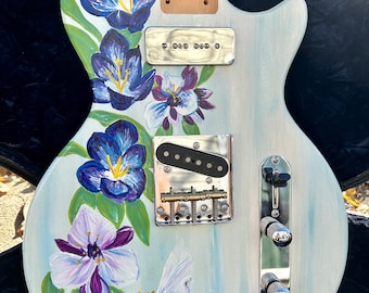 Op de afbeelding: Een witte elektrische gitaar met een bloemenmotief dat op de body is geschilderd. De bloemen zijn blauw, paars en wit. De gitaar heeft een chromen brug en chromen knoppen.