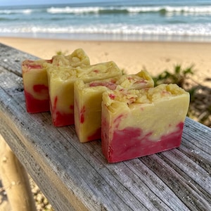 May include: Four handmade soap bars with a layered design. The bottom half of each bar is a vibrant pink, while the top half is a creamy yellow. The soaps are arranged on a weathered wooden surface, with a beach and ocean in the background.