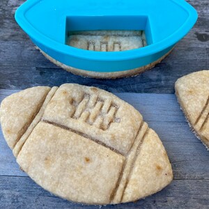 以下が含まれることがあります： フットボールの形をしたシュガー クッキーが 3 つあり、2 つは完全に焼けていて、1 つは青いプラスチック製のフットボール クッキー カッターで部分的に焼かれています。クッキーには茶色の砂糖の釉薬とフットボールのシームのデザインが施されています。