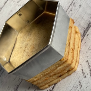 Może przedstawiać: Srebrna sześciokątna forma do pieczenia leży na stosie czterech jasnobrązowych ciasteczek.