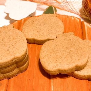 May include: Four pumpkin-shaped sugar cookies on a wooden surface. The cookies are a light brown color and have a slightly rough texture.