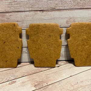 May include: Three brown sugar cookies shaped like coffee cups. The cookies are on a light brown wooden surface.