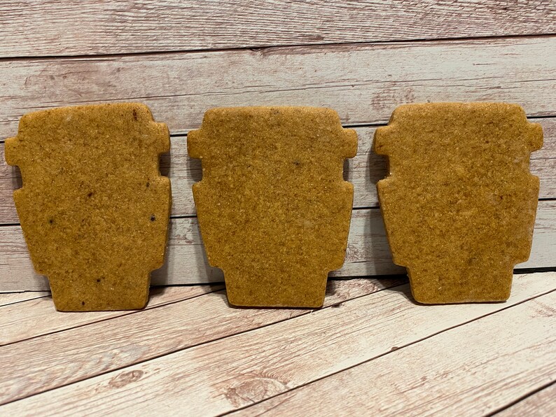 May include: Three brown sugar cookies shaped like coffee cups. The cookies are on a light brown wooden surface.