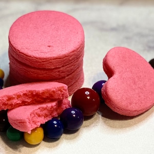 May include: A stack of pink sugar cookies, one broken in half, and a pink heart-shaped cookie. The cookies are surrounded by colourful beads.
