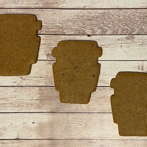 May include: Three brown gingerbread cookies shaped like coffee cups. The cookies are on a white wooden surface.