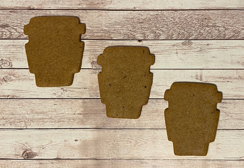 May include: Three brown gingerbread cookies shaped like coffee cups. The cookies are on a white wooden surface.