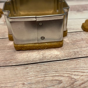 May include: A silver metal cookie cutter shaped like a house, with two small rivets on the side, is placed on top of a brown gingerbread cookie.