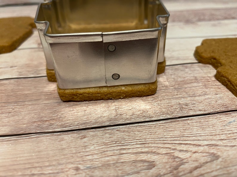 May include: A silver metal cookie cutter shaped like a house, with two small rivets on the side, is placed on top of a brown gingerbread cookie.