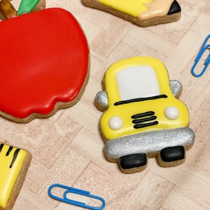 May include: A set of four decorated sugar cookies, including a red apple, a yellow school bus, a yellow ruler, and a yellow pencil. The cookies are decorated with icing and are perfect for a back-to-school party.